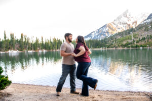 Man and woman hold hands and dance while smiling on beach in front of lake and mountains, captured by Grand Teton engagement photographer.