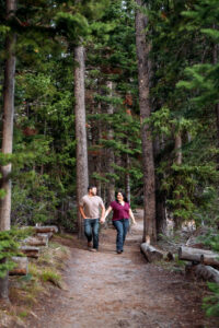Man and woman holding hands and smiling at each other as they walk on trail lined with tall, green trees, captured by Grand Teton engagement photographer.