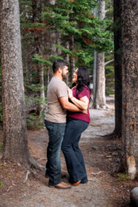 Man and woman facing each other and smiling with woman's hands on man's chest, framed by trees on trail, captured by Grand Teton engagement photographer.