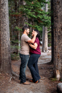 Man and woman facing each other as man brushes woman's hair from her face, framed by trees on trail, captured by Grand Teton engagement photographer.