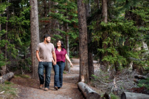 Man and woman holding hands and smiling as they walk on trail lined with tall, green trees, captured by Grand Teton engagement photographer.