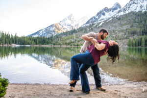 Man dipping woman and woman laughing on beach in front of lake and mountains, captured by Grand Teton engagement photographer.