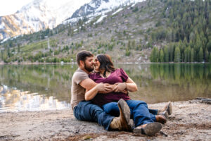 Man sitting on beach with woman leaning against him and the two touching noses in front of river and mountains, captured by Grand Teton engagement photographer.
