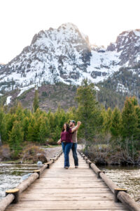 Man and woman smile and hold hands and extend arms on bridge over river with mountains in back, captured by Grand Teton Engagement photographer.