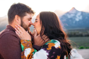 Man and woman's hands cupping each other's faces while their noses touch and they smile in front of mountains at sunset, captured by Grand Teton engagement photographer.