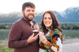 Man and woman facing each other and smiling at camera while holding two cupcakes with a topper in front of mountains, captured by Grand Teton engagement photographer.