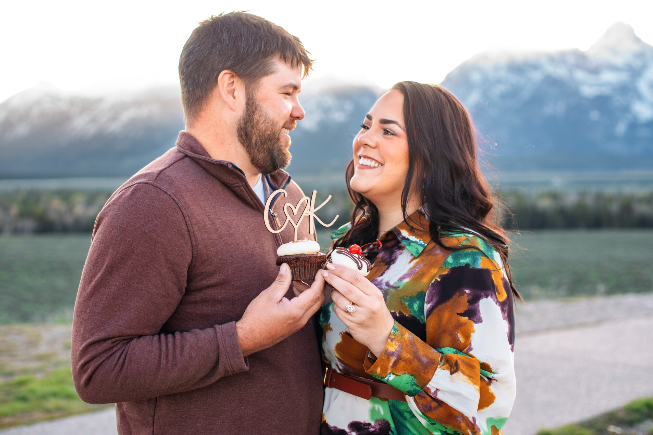 Man and woman smiling at each other while holding two cupcakes with a topper in front of mountains, captured by Grand Teton engagement photographer.