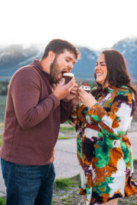 Man opening mouth to eat cupcake while woman laughs in front of mountains, captured by Grand Teton engagement photographer.