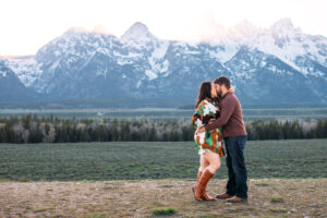 Man and woman kissing with man's hand on woman's hip and woman wrapping her arm around man's neck in front of mountain at sunset, captured by Grand Teton engagement photographer.