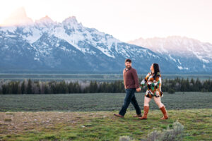 Man and woman holding hands and smiling at each other as they walk across grass in front of mountains at sunset, captured by Grand Teton engagement photographer.
