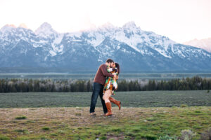 Man holding and kissing woman as woman places hand on man's neck and pops a foot in front of mountains at sunset, captured by Grand Teton engagement photographer.