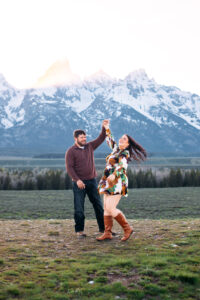 Mand and woman dancing, with one arm held high and woman flipping hair in front of mountains at sunset, captured by Grand Teton engagement photographer.