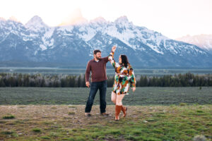 Man and woman dancing with hands held and an arm raised in front of mountains at sunset, captured by Grand Teton engagement photographer.