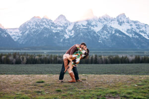 Man dipping and kissing woman as he holds her thigh in front of mountains at sunset, captured by Grand Teton engagement photographer.