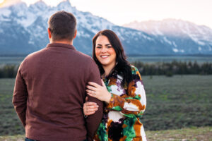 Man facing mountains as woman faces and smiles at camera with hands resting on man's arm during sunset, captured by Grand Teton engagement photographer.