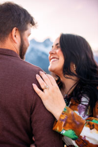 Woman's wedding ring set on hand that rest's on man's shoulder and woman smiling up at man in front of mountains at sunset, captured by Grand Teton engagement photographer.