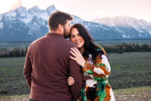 Man facing mountains and woman facing and smiling at camera as she rests head on man's shoulders and man kisses her forehead in front of mountains at sunset, captured by Grand Teton engagement photographer.