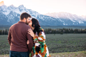 Man facing mountains as women faces camera and kisses him on lips in front of mountains at sunset, captured by Grand Teton engagement photographer.