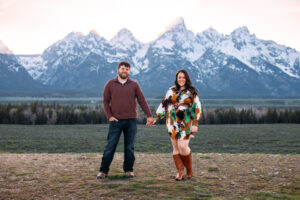 Man with one hand in pocket holds hands with woman and they both smiles at camera in front of mountains at sunset, captured by Grand Teton engagement photographer.