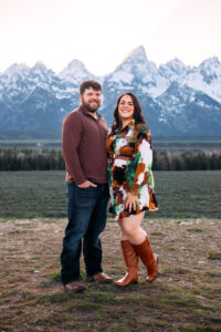 Man with one hand in pocket angles to women with hands resting on thigh and they both smile at camera in front of mountains at sunset, captured by Grand Teton engagement photographer.