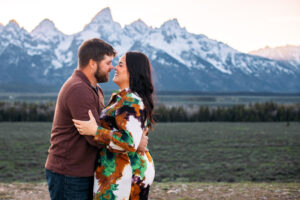 Man and woman face each other and hold each other as they touch noses and smile in front of mountains at sunset, captured by Grand Teton engagement photographer.