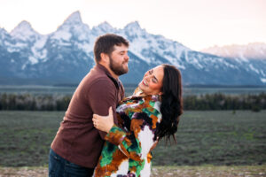 Man holds woman as woman leans back and smiles in front of mountains at sunset, captured by Grand Teton engagement photographer.