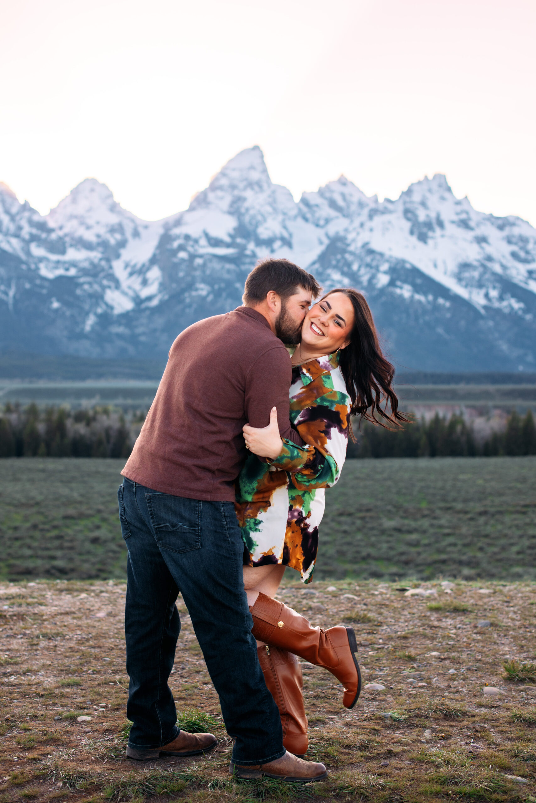 Man holds woman and buries his face in her neck as woman leans back, pops her foot, smiles at camera, and flips hair in front of mountains at sunset, captured by Grand Teton engagement photographer.