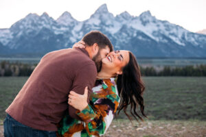Man holds woman and presses his face into her neck as woman leans back and laughs in front of mountains at sunset, captured by Grand Teton engagement photographer.
