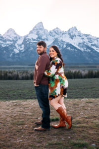 Man stands with hands in pockets as woman hugs him from behind and pops her foot and they both smile at the camera in front of mountains at sunset, captured by Grand Teton engagement photographer.
