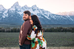 Man stands with hands in pockets and smiles at camera as woman hugs him from behind and stares off into the distance in front of mountains at sunset, captured by Grand Teton engagement photographer.