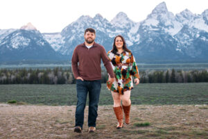 Man and woman smile and walk towards camera with man leading and placing one hand in pocket in front of mountains at sunset, captured by Grand Teton engagement photographer.