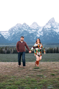 Man and woman smiling and walking towards camera with man placing one hand in pocket in front of mountains at sunset, captured by Grand Teton engagement photographer.