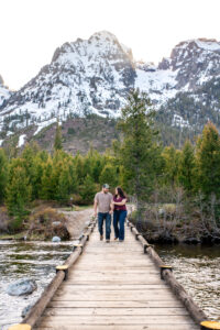 Man and woman smile at each other and walk as woman hold man's arm and man puts hand in pocket on bridge over river with mountains in back, captured by Grand Teton Engagement photographer.