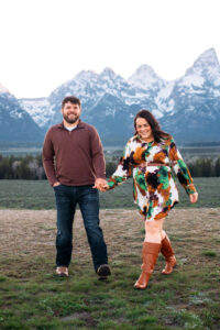 Man and woman smiling an walking towards camera as woman looks down in front of mountains at sunset, captured by Grand Teton engagement photographer.