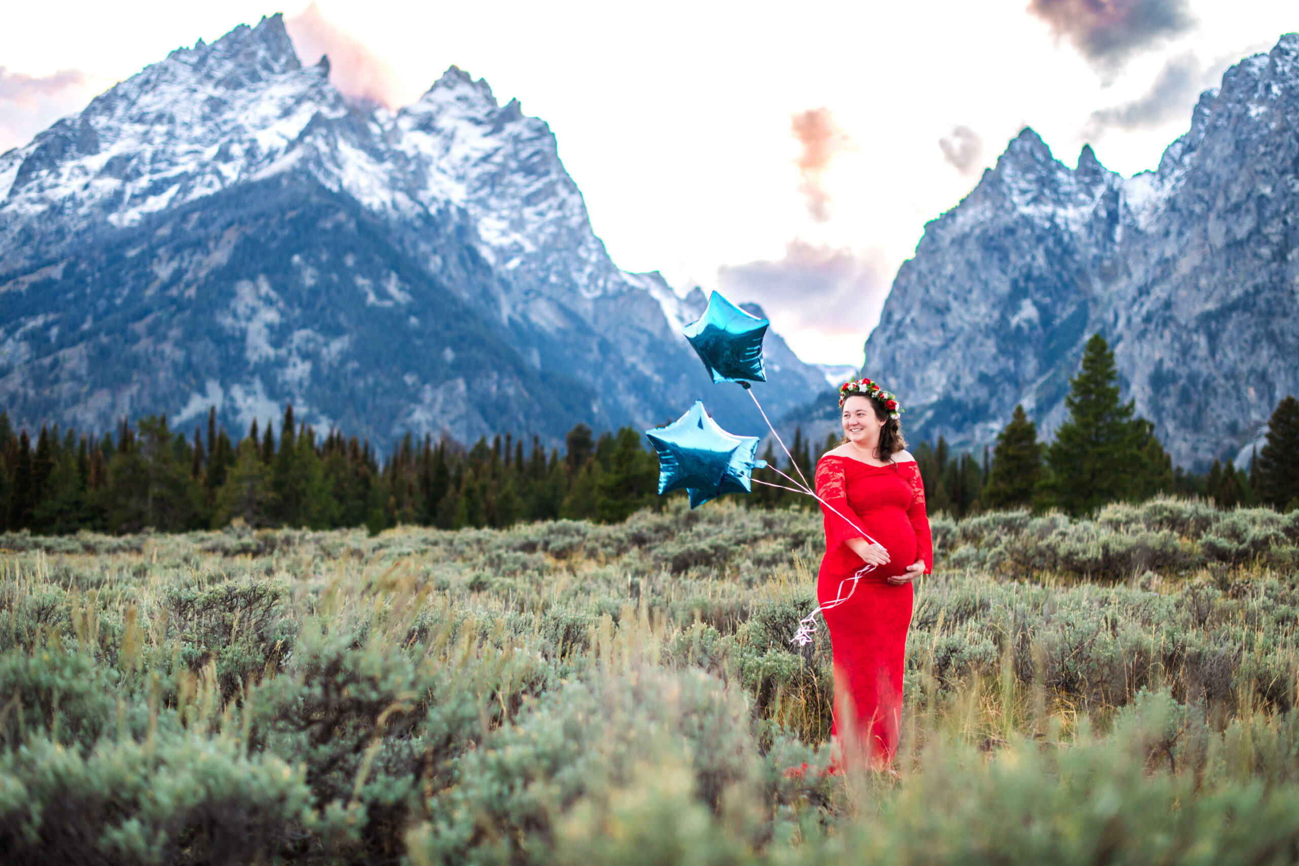 Woman in red dress plays with blue balloons and holds baby bump in front of mountains, captured by Grand Tetons maternity photographer.