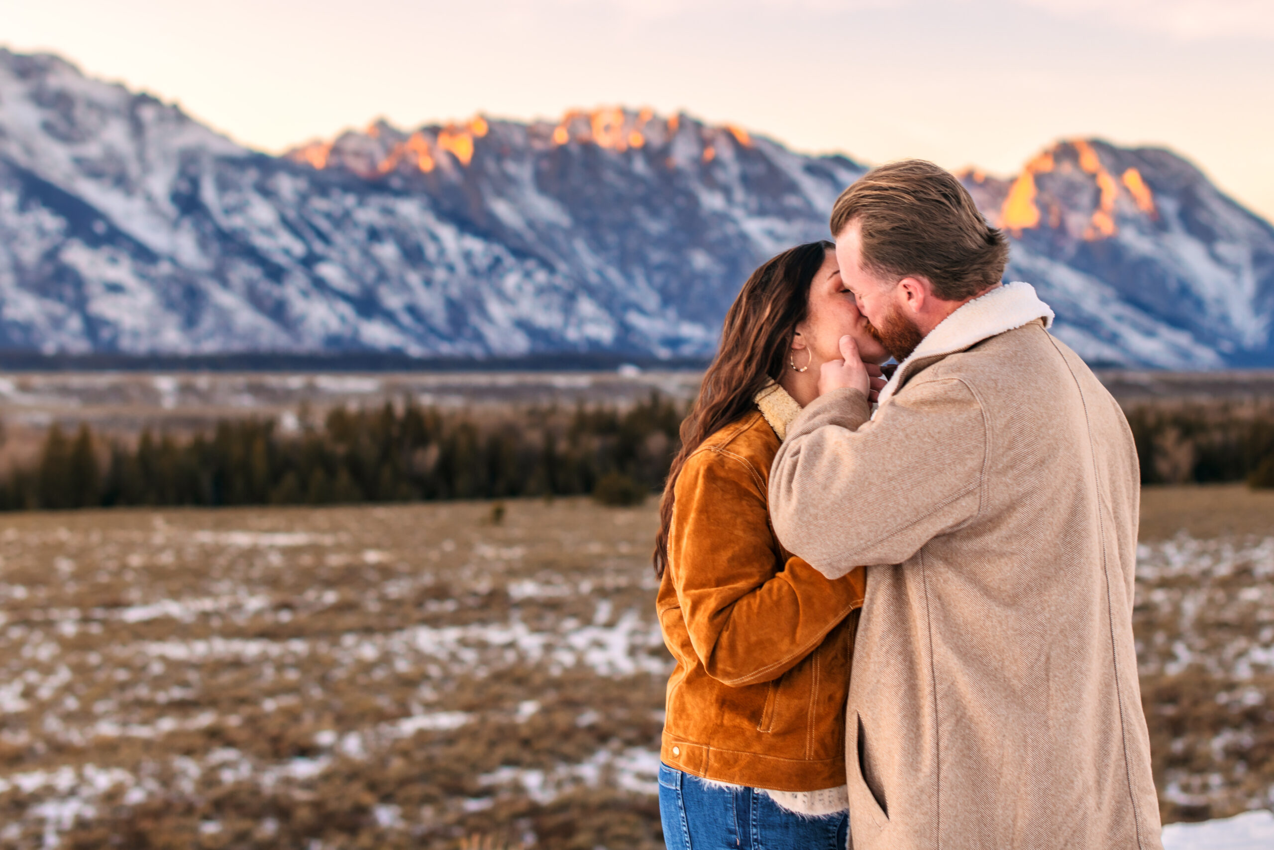 Husband gripping wife's jaw and kissing her in front of mountain range with sunset alpenglow, capture by Grand Teton National Park photographer.
