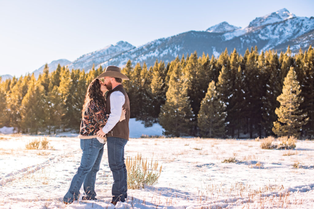 Husband and wife kiss in winter landscape in front of green, snow covered trees in Grand Teton National Park.