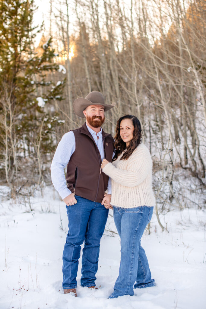 Man with hand in pocket smiles at camera with wife holding his arm in front of aspen trees at Grand Teton National Park.