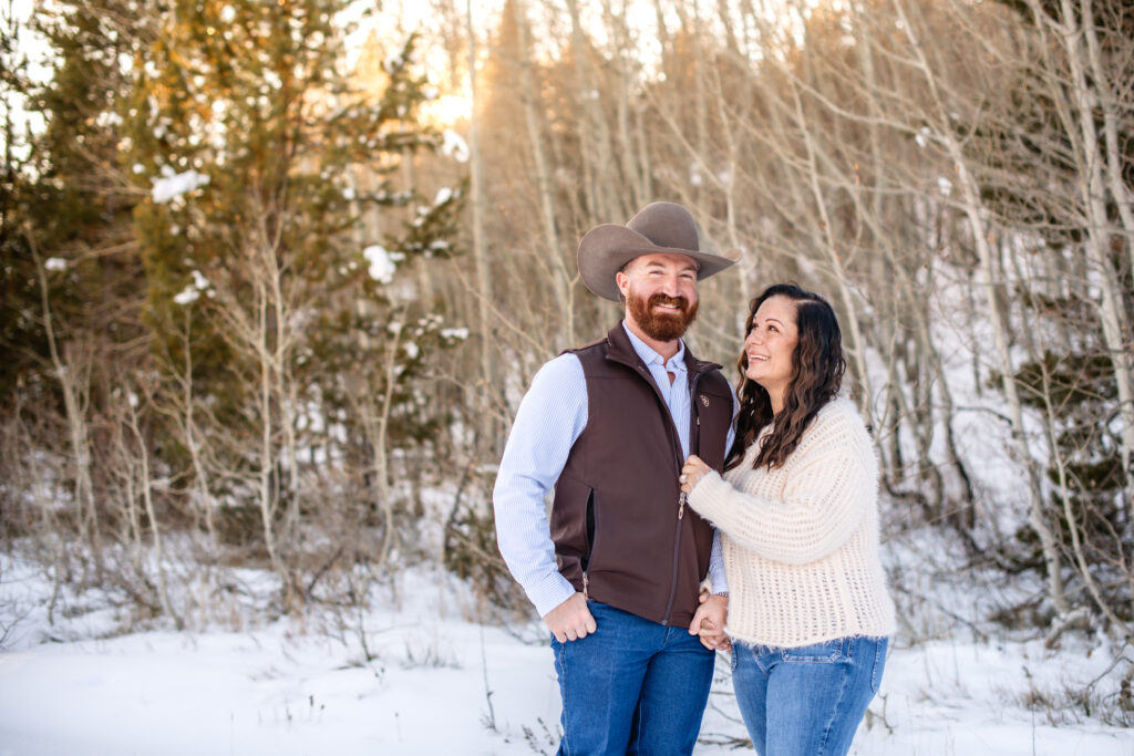 Husband with cowboy hat and hands in pocket smiles at camera as wife smiles up at husband in front of wintery landscapes with aspens and the sun peeking through at Grand Teton National Park.