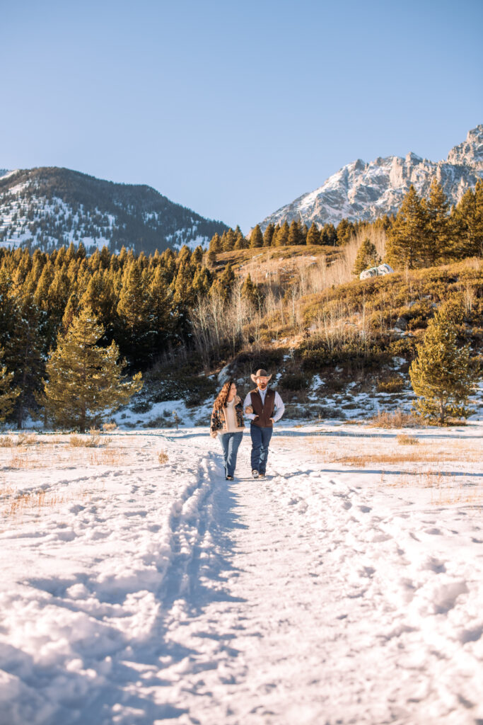 Man with cowboy hat and woman link arms and walk towards the camera in the snow in front of the Grand Tetons.