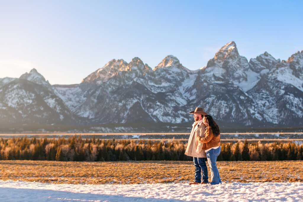 Wife hugs husband with cowboy hat from behind as they stare out towards the sun in front of the Grand Teton mountain range.