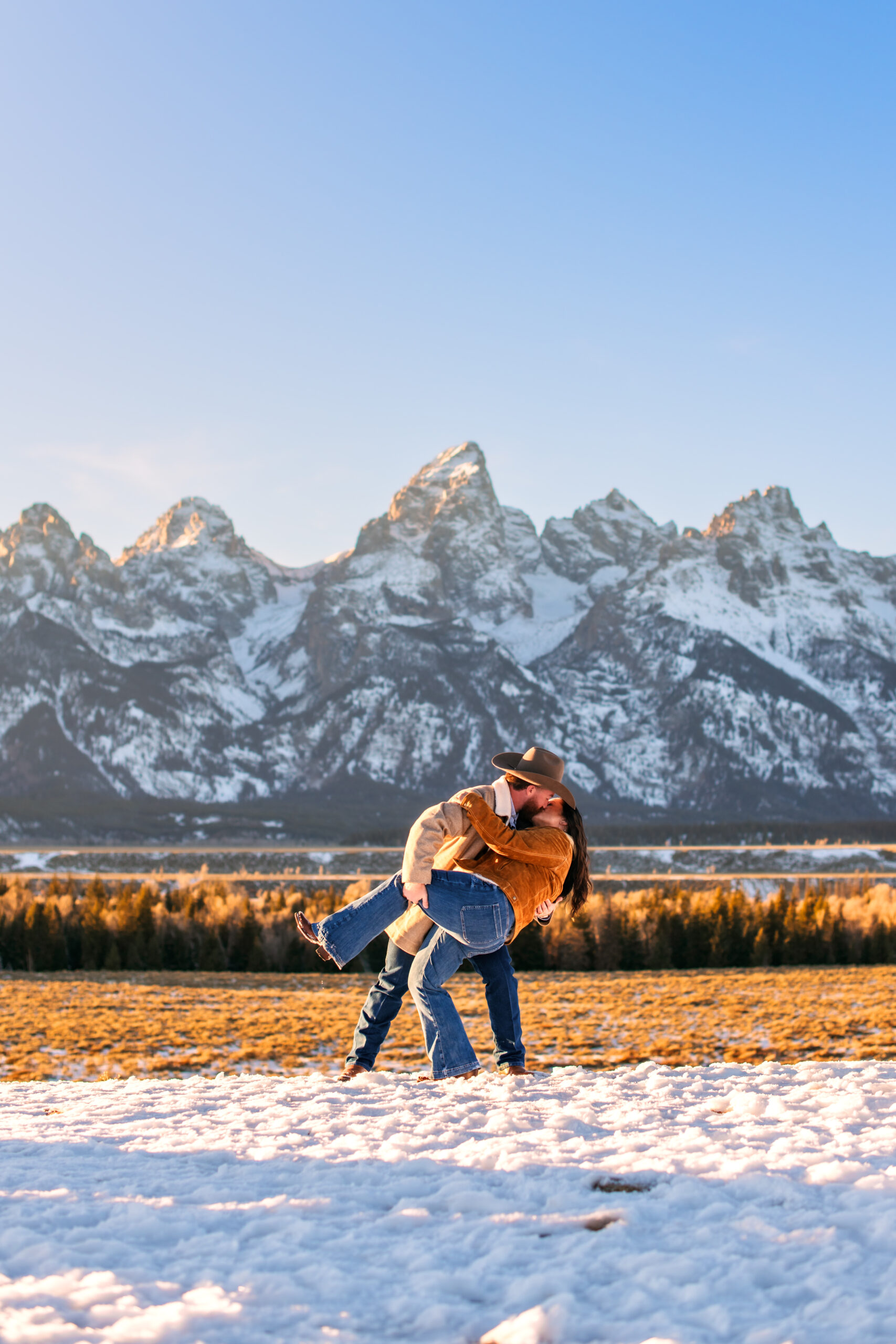 Husband with cowboy hat holds wife's thigh as he dips and kisses her romantically in wintery landscape in front of mountain range, captured by Grand Teton National Park photographer.