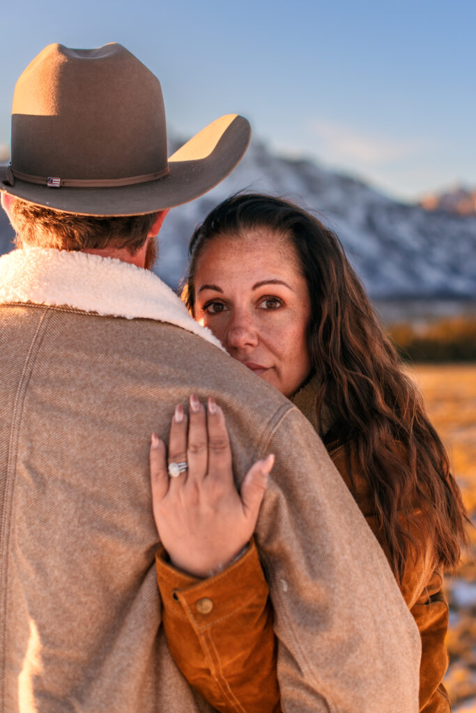Wife staring at camera over husband's shoulder with her hand and wedding ring on his back in front of the Grand Tetons.