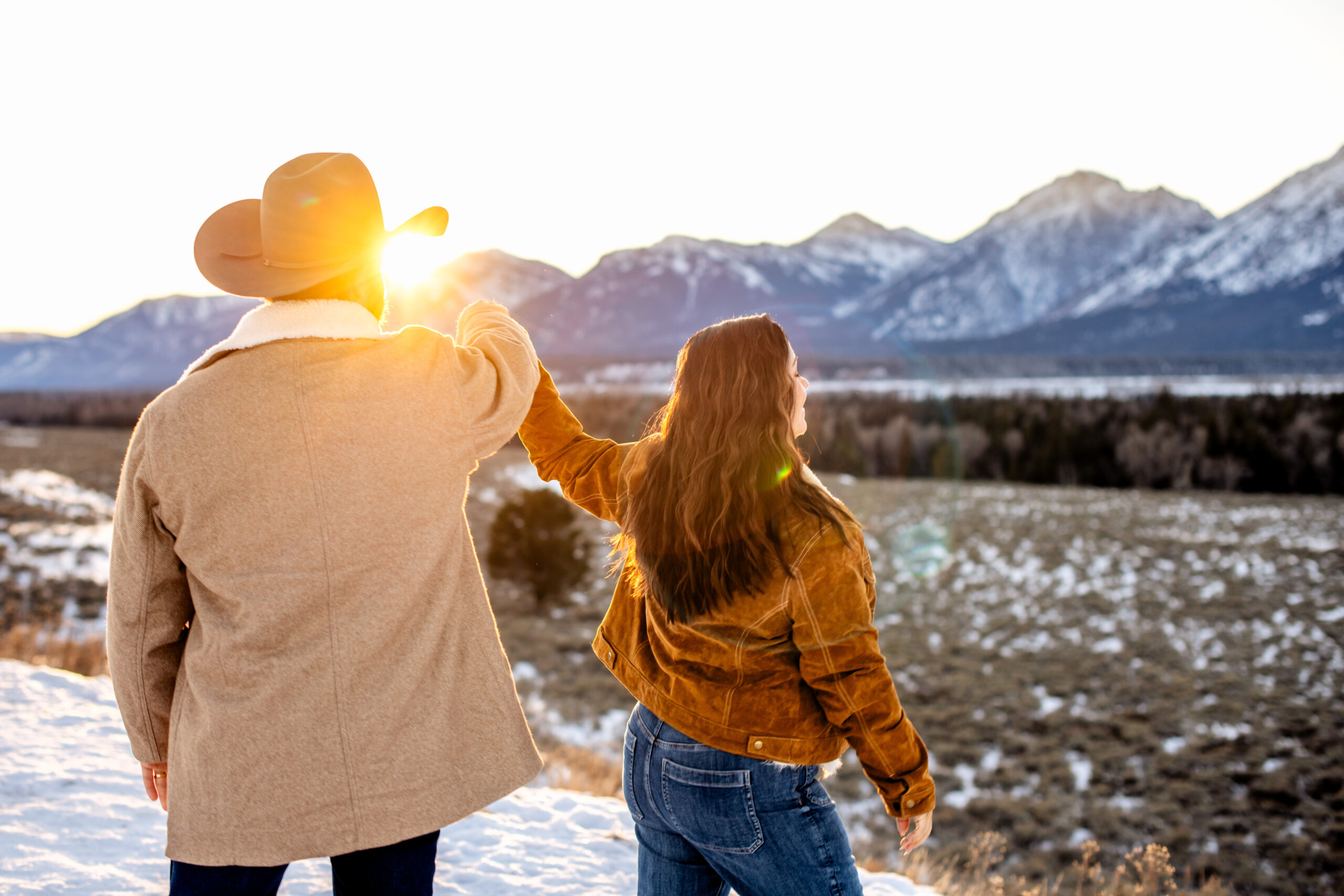 Husband with cowboy hat twirling wife, who smiles, as sunset flares between them in snowy mountain landscape, captured by Grand Teton National Park photographer.
