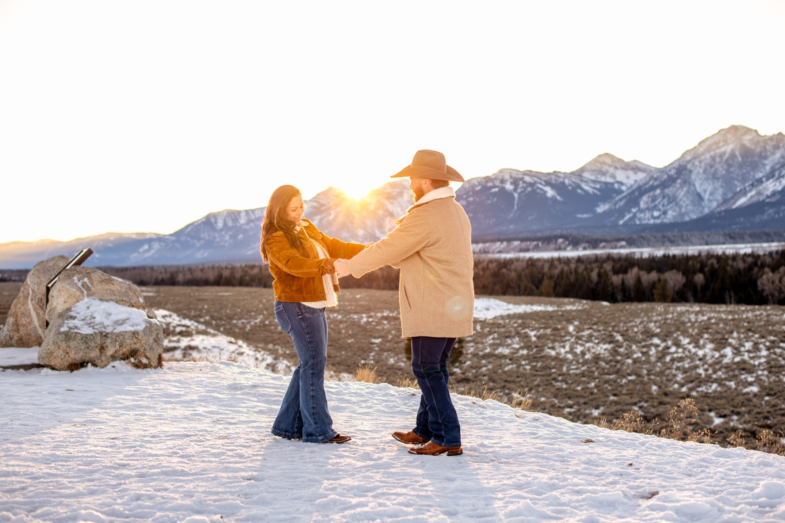 Husband and wife hold hands and dance in front of mountains on snowy landscape with sun flare, captured by Grand Tetons engagement photographer.