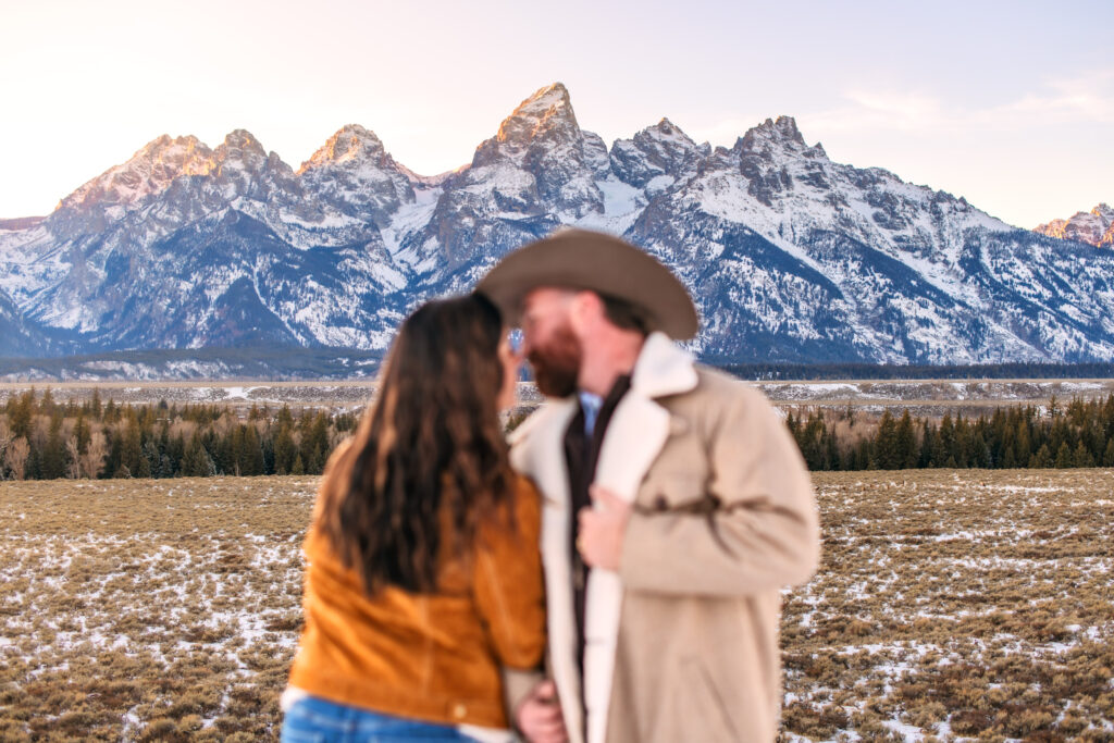 Husband and wife are out of focus as camera focuses on the Grand Teton Mountain range during sunset.