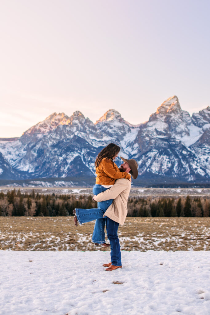Husband lifts wife and they smile at each other as wife pops her leg in winter landscape in front of the Grand Tetons.