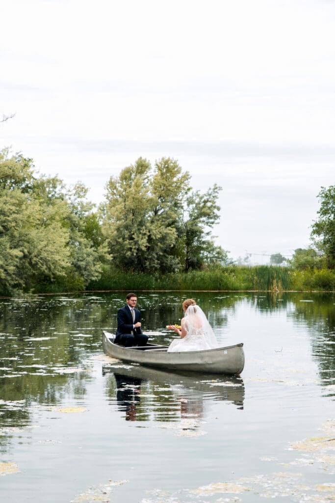 Groom paddles in row boat as bride offer charcuterie board on lake at the Snake River Meadow wedding venue, captured by Idaho Falls wedding photographer.