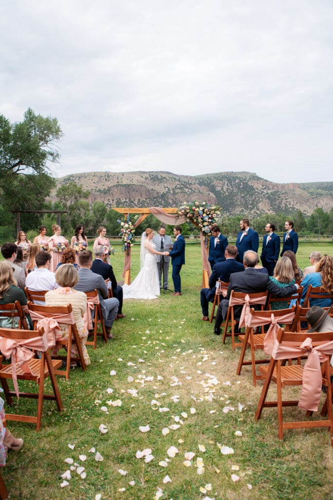 Bride and groom hold hands in front of arch and officiant with family and friends watching with hills in background at the Snake River Meadow wedding venue behind them, captured by Idaho Falls wedding photographer.