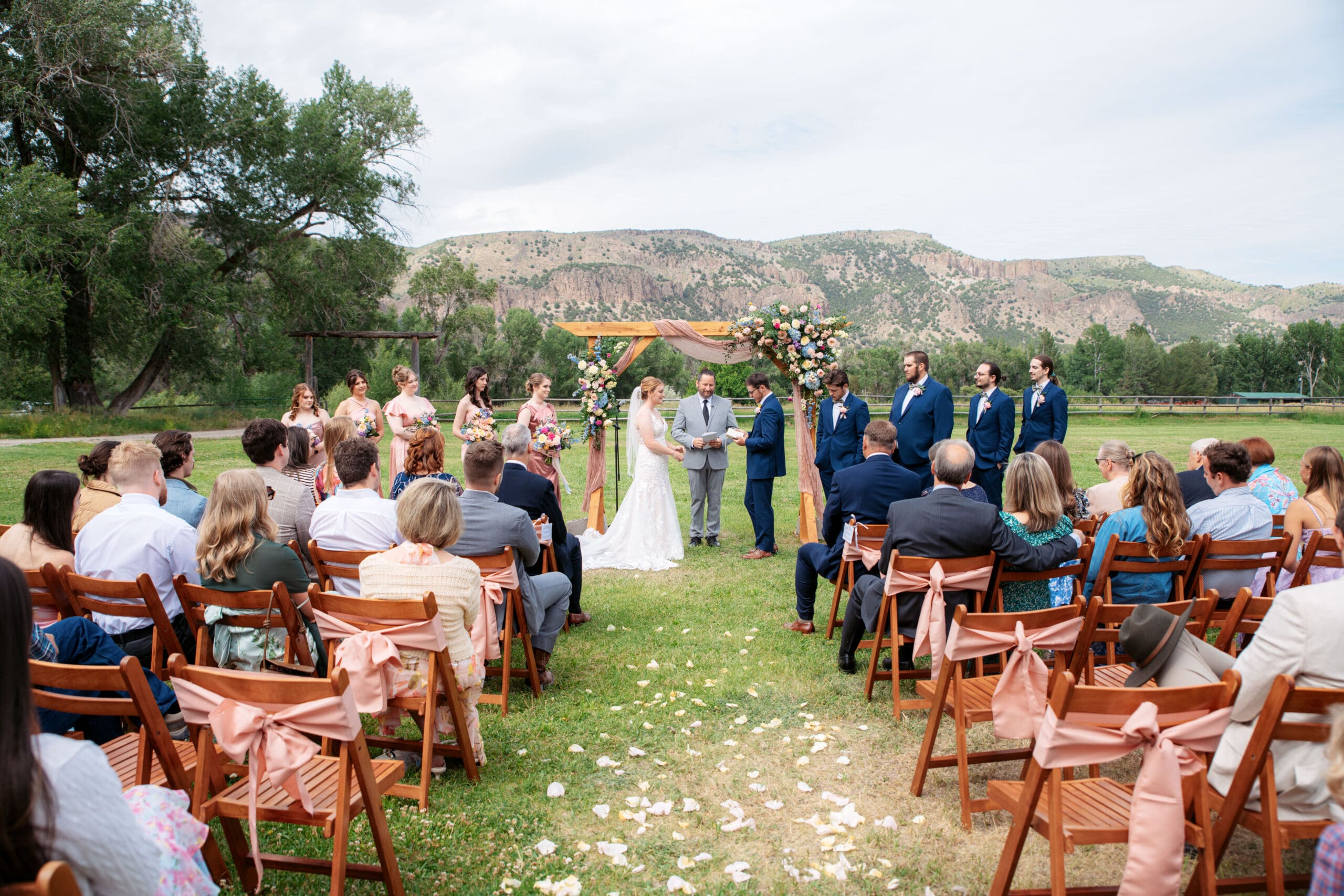Bride and groom hold hands in front of arch and officiant with groomsmen and bridesmaids lined on sides and family and friends watching with hills in background at the Snake River Meadow wedding venue behind them, captured by Idaho Falls wedding photographer.